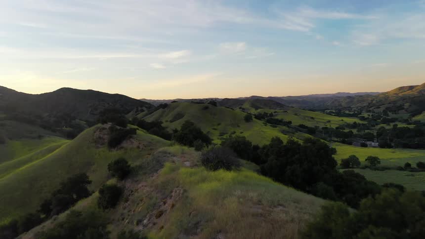 An aerial shot shows a vast landscape of rolling green hills and scattered trees under a clear sky as the sun begins to set