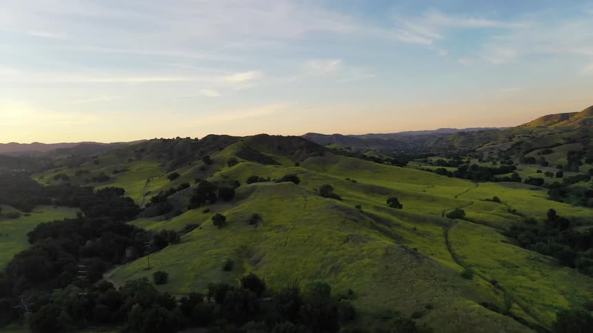 An aerial shot sweeps over rolling green hills and dark silhouetted mountains during a quiet dusk