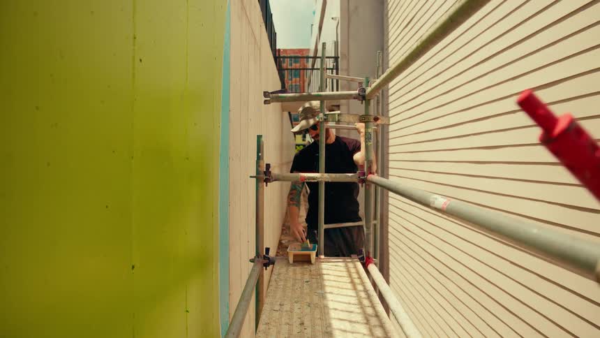 A man in a hat stands on scaffolding and paints a vibrant limegreen mural on an outdoor wall looking forward with focus