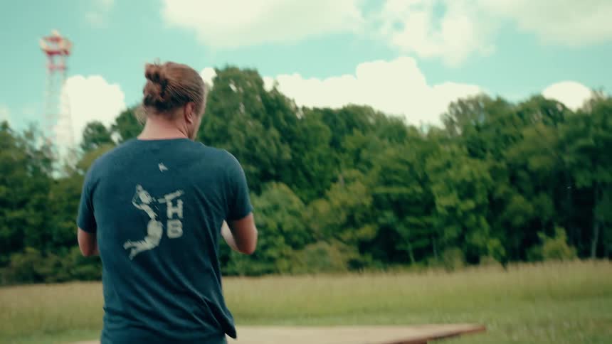 A man with a bun in a dark tshirt places a black box on a wooden table then he open it in a grassy field with trees in the background