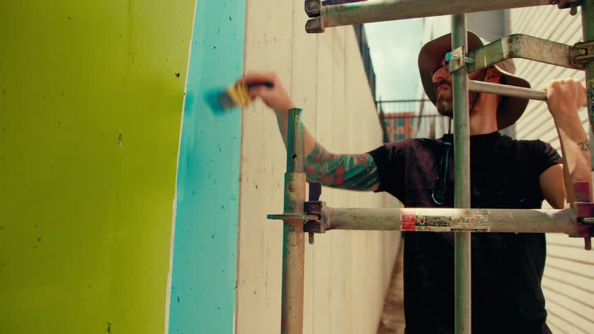 A medium shot shows a man with arm tattoos and a sun hat standing on scaffolding as he meticulously paints a blue section of a large outdoor mural