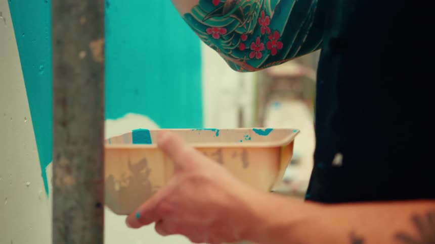 A closeup shot shows a man with tattoos and a bucket hat painting a vibrant blue mural on a city wall