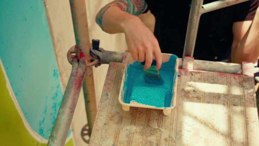 Person applying blue paint to vertical wall with brush and container on scaffold
