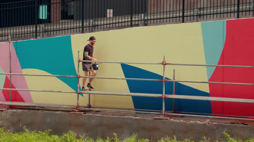 A wide shot shows a man with tattoos walking along scaffolding in front of a large multicolored geometric mural on a long city wall