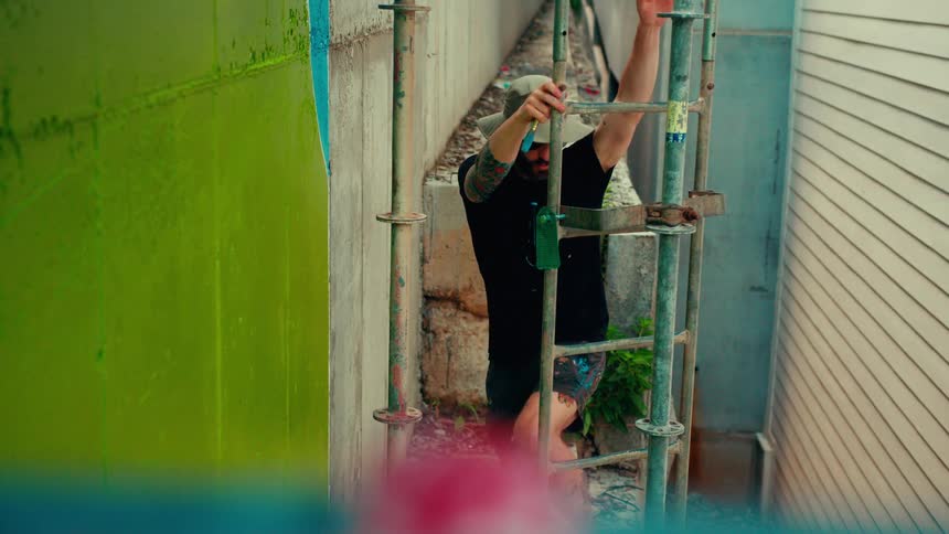 A man in a tan hat stands on scaffolding grabbing paint before painting a bright green mural on a concrete wall