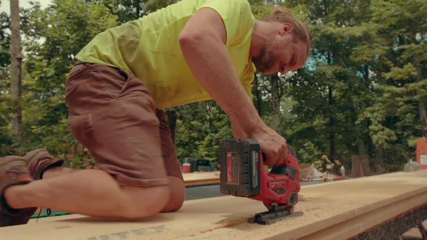 A worker in a yellow shirt and brown shorts kneels on a large panel while using a red jigsaw to cut through the material at a construction site