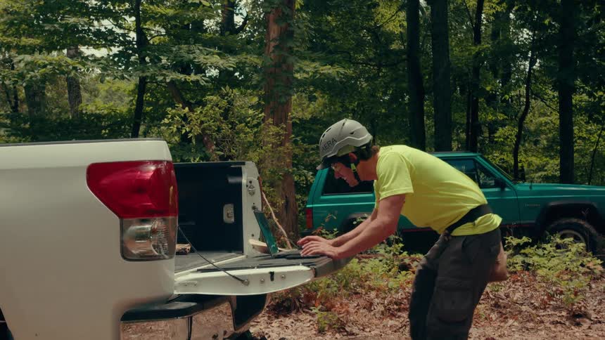 A construction worker wearing a helmet stands behind his truck in the woods at midday using his laptop as sunlight filters through the trees