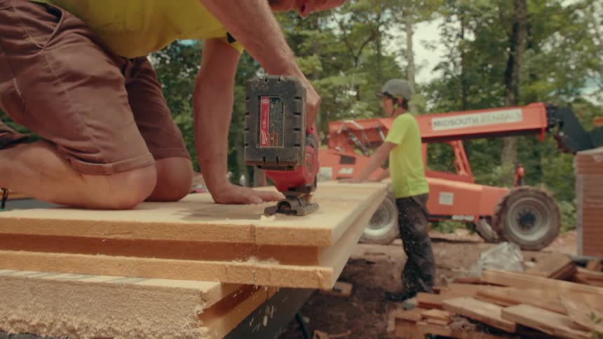 A construction worker kneels on a stack of boards and uses a jigsaw to cut through them while another worker stands by a telehandler in a wooded construction site