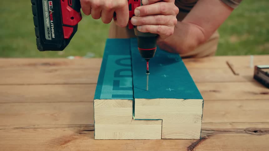A man uses a power drill to drive a long screw into a tealpainted wooden beam outdoors