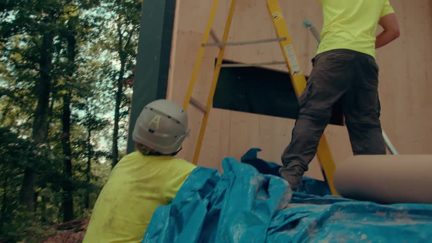 A man in a limegreen shirt climbs a ladder and a man in a grey helmet work together to secure a wooden wall frame in a forest
