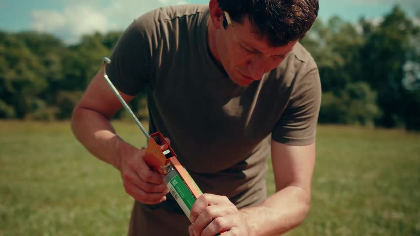 A closeup shot shows a man in a brown tshirt meticulously applying a line of green adhesive from a caulking gun onto a piece of blue construction material outdoors