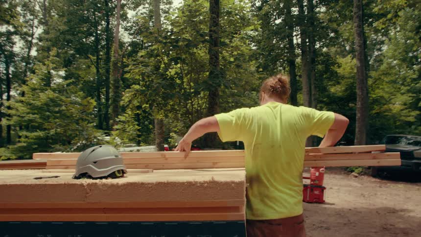 A man in a limegreen shirt carries three long wooden planks toward a construction site in a forest