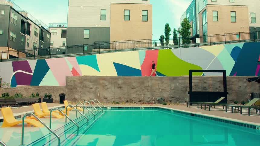 A man in a black shirt and tan hat walks along a ledge beside a colorful geometric mural overlooking a blue swimming pool