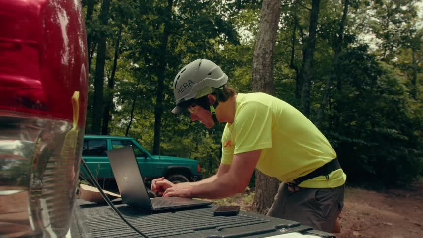 A man wearing a safety helmet and a yellow shirt stands at the tailgate of a white pickup truck using a laptop and taking notes while working in a wooded outdoor area next to a blue car
