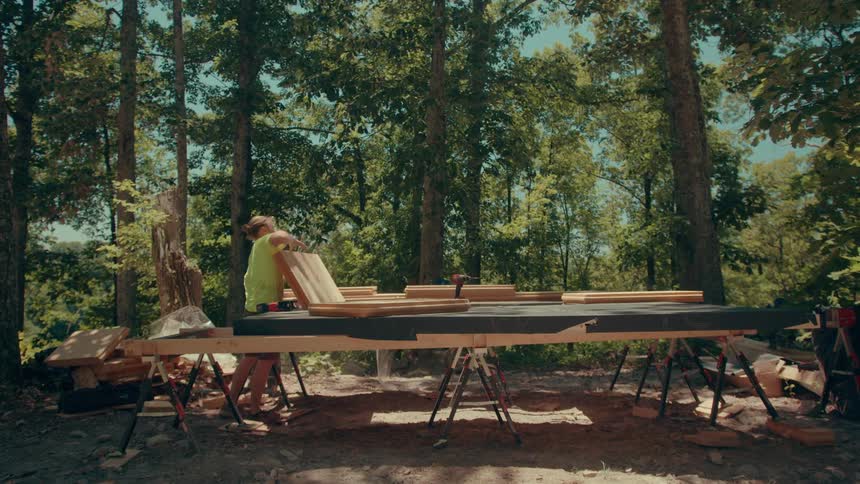 A man in a limegreen shirt and a man in a lightyellow shirt build a wooden structure in a forest looking forward with concentration