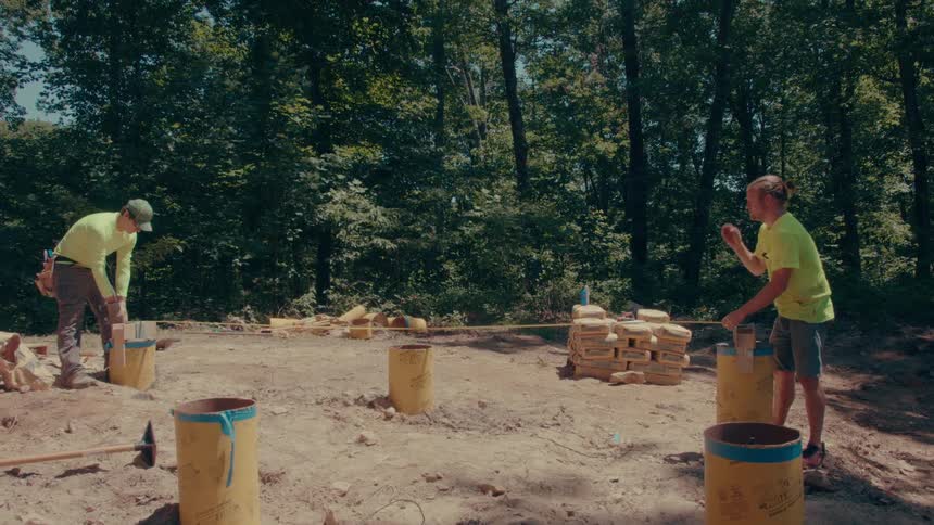Two construction workers in highvisibility shirts use a tape measure across yellow concrete pier forms to check the dimensions of a building foundation on a cleared dirt site surrounded by woods