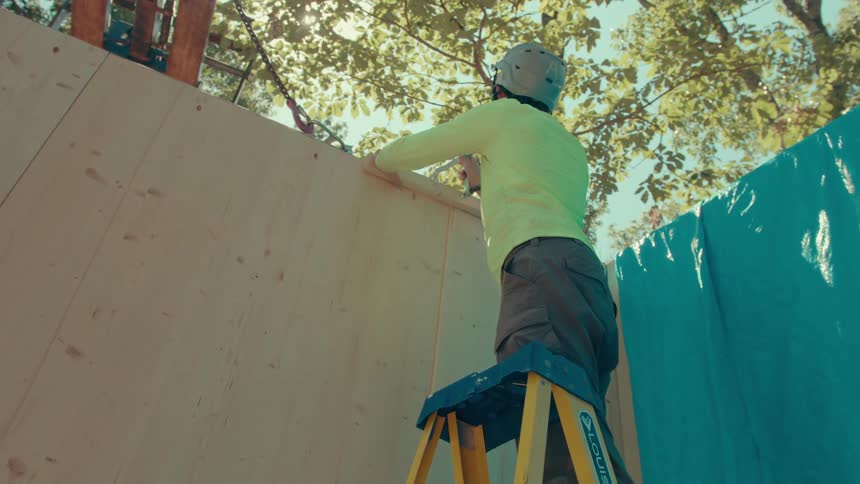 A man in a limegreen shirt and grey helmet stands on a yellow ladder as a man below hands him wood to position the wooden beam against a wall in a forest