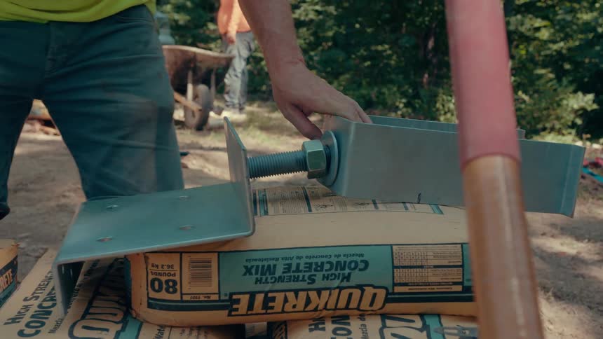 A construction worker wearing a yellow shirt and work shorts leans over several bags of highstrength concrete to assemble and adjust a large metal bracket and bolt system at an outdoor job site