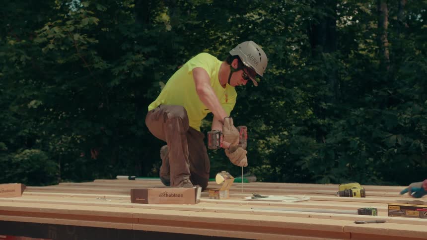 Worker kneeling with drill on wooden platform in forested area with tools around