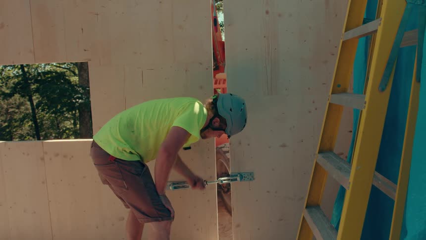 A man in a grey helmet and limegreen shirt uses a silver tool to tighten a metal latch on a wooden wall