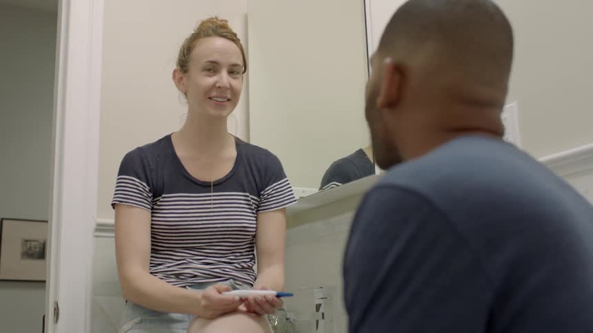 A woman holding a pregnancy test talks to a man in the bathroom