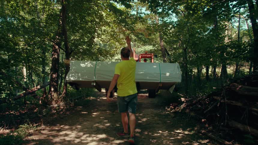 A man in a limegreen shirt walks with his arms raised while guiding a tractor carrying a large white load through a forest looking forward with focused energy