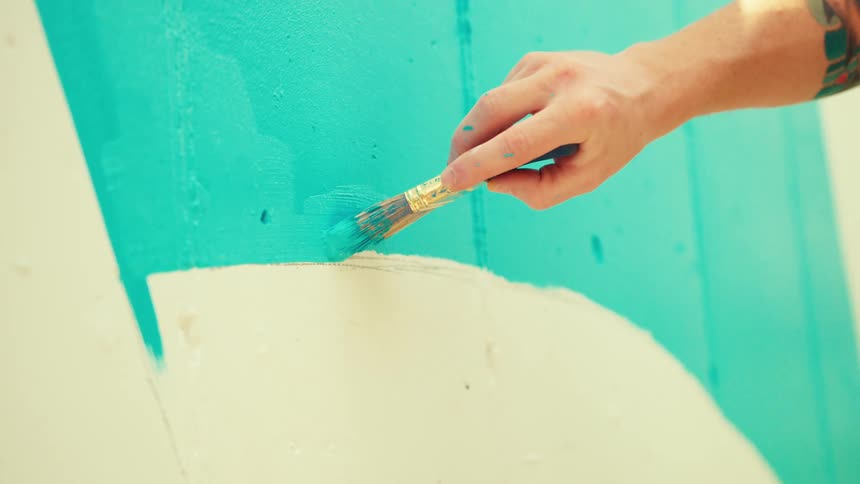 A person with a tattooed arm carefully applies bright blue paint to a textured wall using a paintbrush following a curved line marked by tape to create a mural