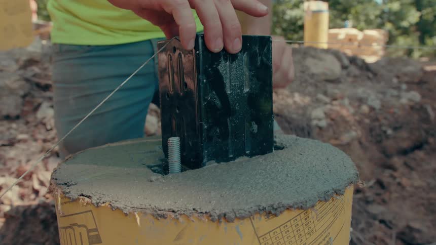 A man in a limegreen shirt uses both hands to secure a black metal post base into wet concrete within a yellow cylindrical form