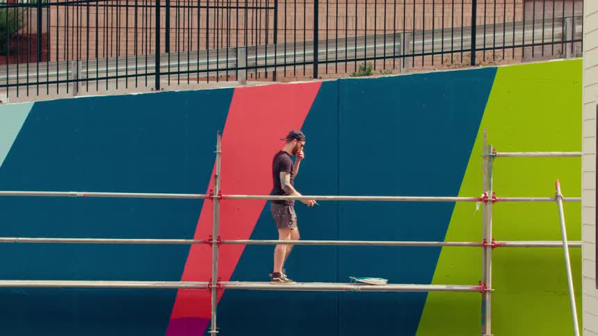 A man in a tan hat walks on scaffolding to grab a paint tray to sit on a ladder as he takes a break from painting on a large outdoor mural