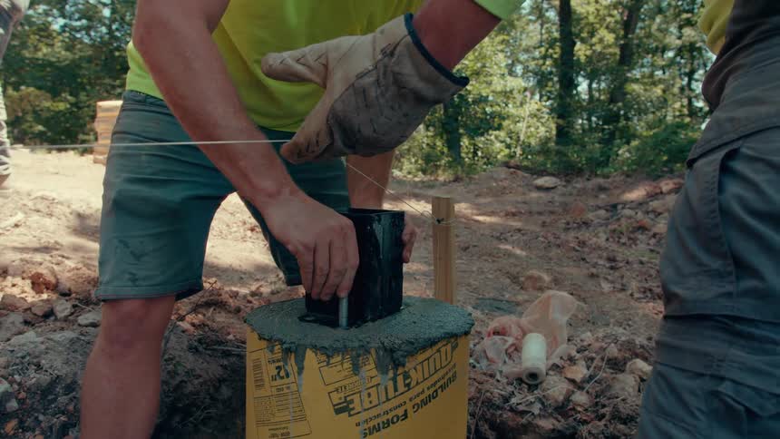 A closeup shot shows a worker in a yellow shirt carefully positioning a black metal bracket into wet concrete within a cylindrical yellow form at a construction site