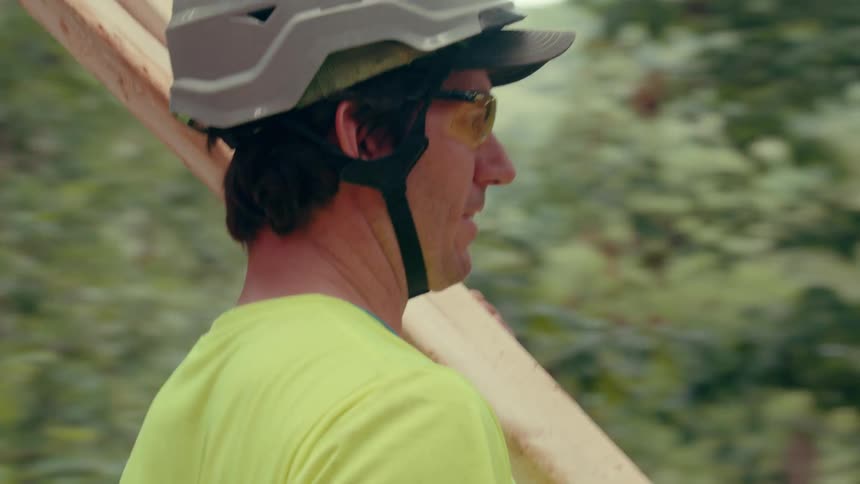 A worker wearing a safety helmet and highvisibility shirt carries a long wooden beam on his shoulder through a wooded construction site