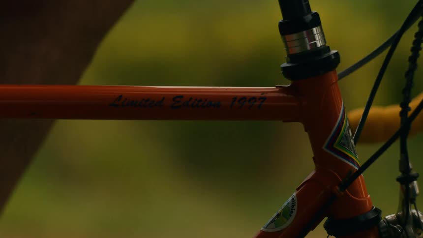 A closeup shot focuses on the orange frame of a bicycle featuring limited edition 1997 text as the person moves in the blurred foreground against a green outdoor setting