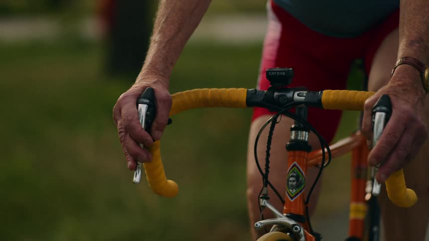 A close up shot shows an elderly man in a blue jersey and red shorts gripping the yellow handlebars of his bicycle while riding outdoors