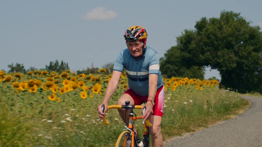 An elderly man in cycling gear rides his bicycle along a paved road past a field of sunflowers during the day