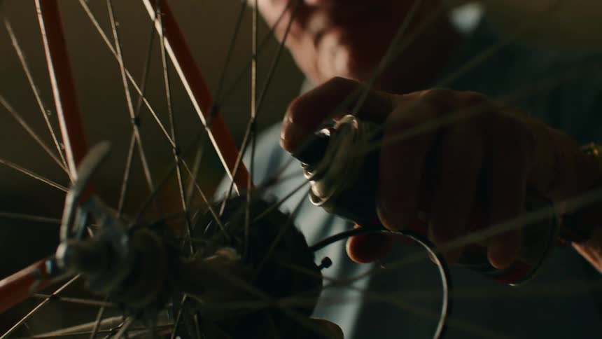 In a dimly lit workshop a man carefully applies lubricant from a spray can to the hub of a bicycle wheel his hands moving with practiced precision among the spokes