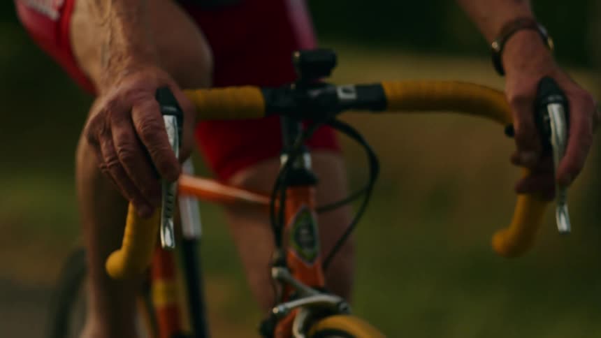 A male cyclist rides a vintage orange road bike along a sunlit path his weathered hands gripping the yellow handlebars as he shifts weight while pedaling in a closeup shot