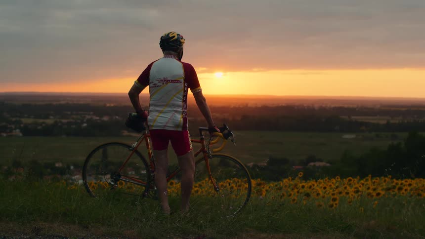 A cyclist in full gear stands with his bike on a grassy hill looking out over a vast field of sunflowers as the sun sets on the horizon