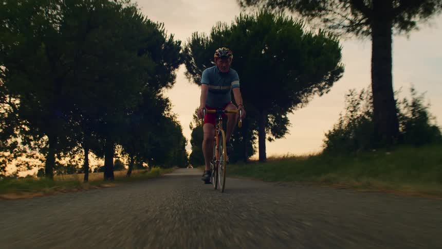 A medium shot shows an elderly man in a blue jersey and red shorts riding his bicycle along a tree lined road at dusk