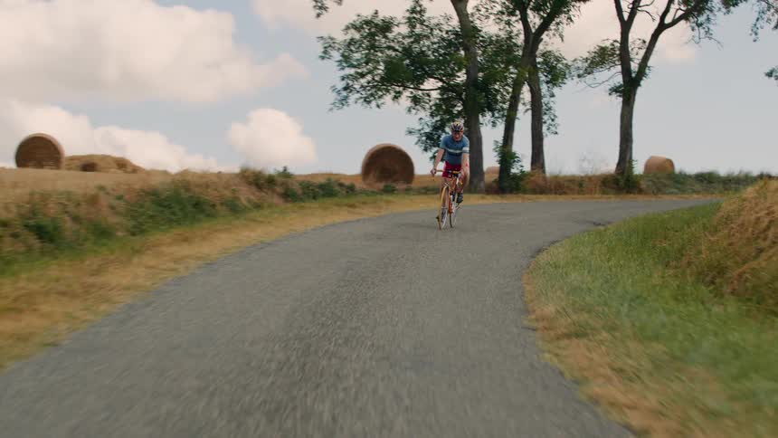 A cyclist pedals at a steady pace up a winding country road with rolling fields dotted with large hay bales visible under a partly cloudy sky