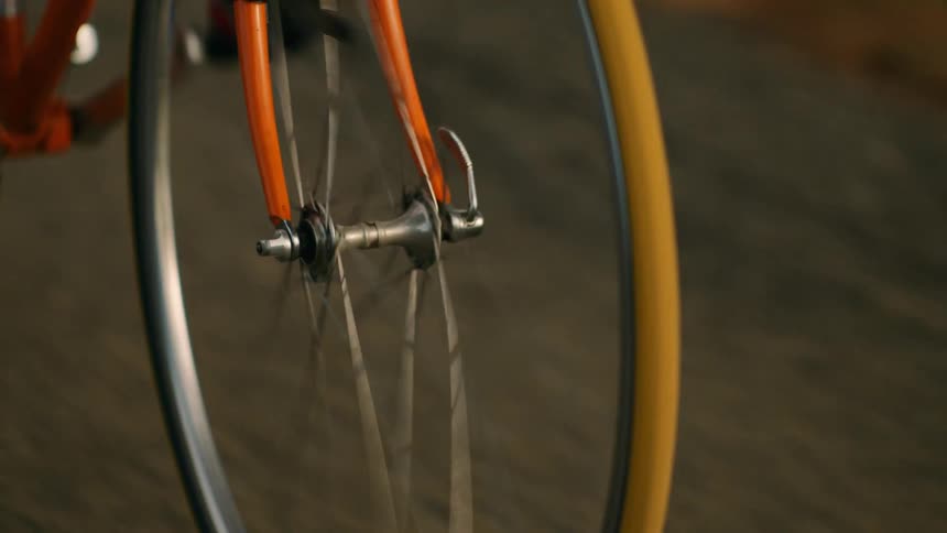 A close up shot shows the spinning front wheel of an orange bicycle as it travels along a paved road
