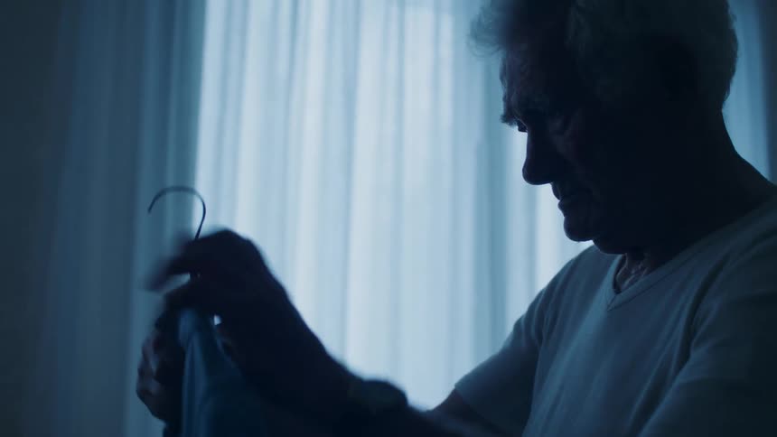 In a dimly lit room an elderly man is seen in silhouette as he picks up a garment on a hanger while standing in front of a window with sheer blue curtains