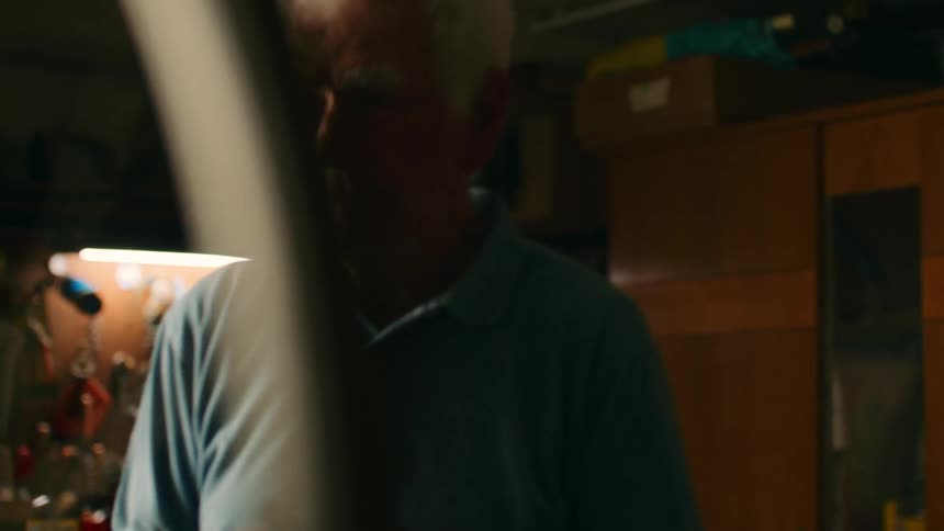 An elderly man with white hair wearing a light blue polo shirt stands in a cluttered workshop before moving to inspect a bicycle wheel