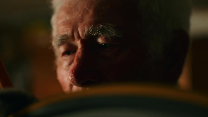 A close up shot focuses on the weathered face of an elderly man with white hair as he intently works on a bicycle in a dimly lit setting