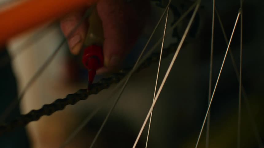 A close up shot shows an older person using a small red bottle to apply oil to a bicycle chain