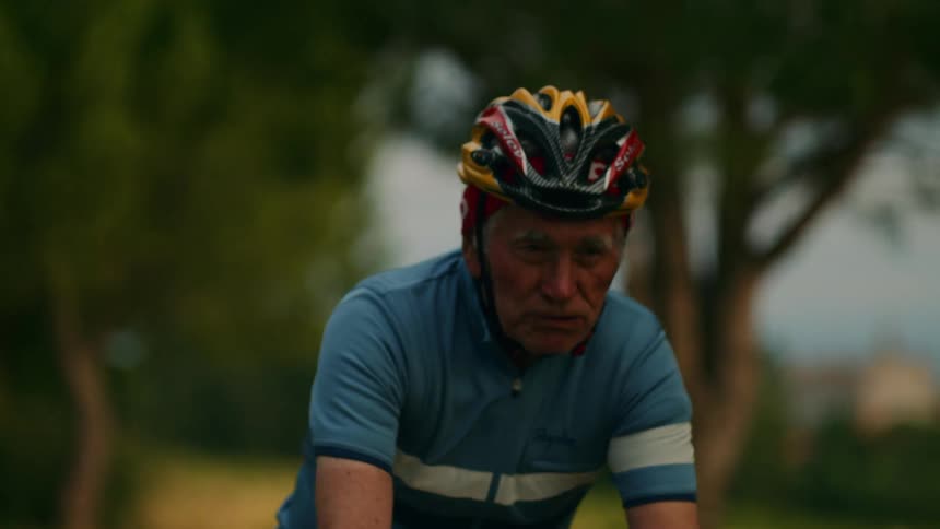 Elderly person cycling outdoors wearing blue jersey and multicolored helmet in wooded area