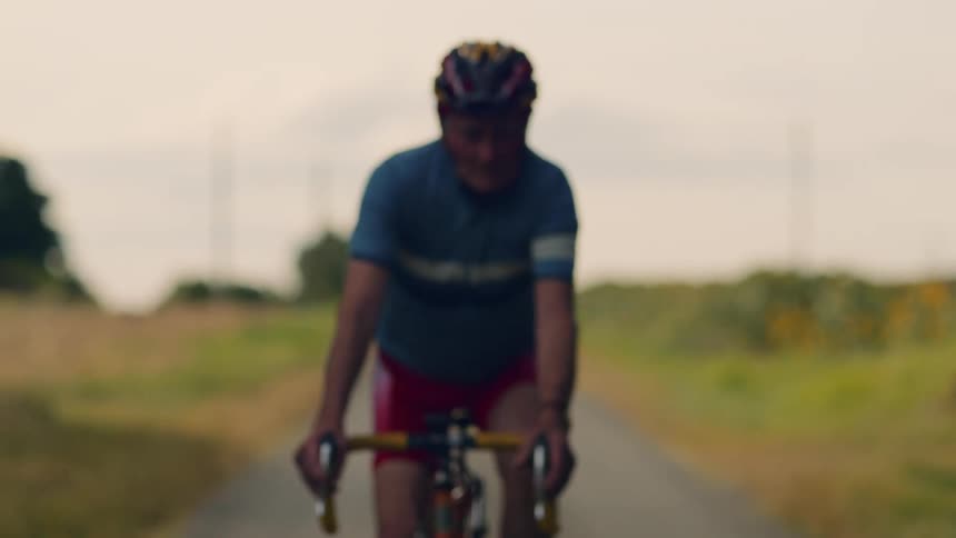 Starting as a softfocus silhouette an older cyclist slowly comes into clear view as he rides with intense determination down a quiet country road