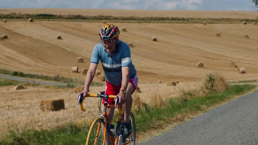 An elderly man wearing professional cycling gear rides his road bike along a paved rural road passing through a harvested wheat field filled with round hay bales under a bright sky
