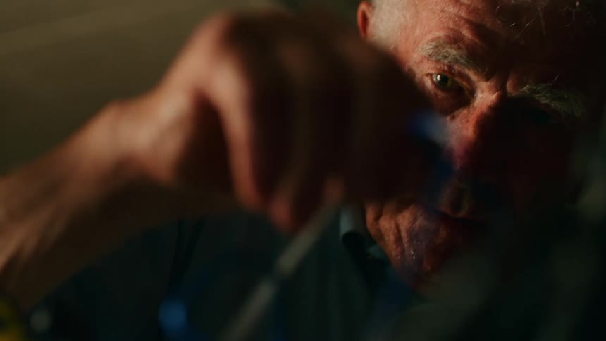 An elderly man focuses intently as he uses an allen key to adjust a blue bicycle frame in a dimly lit workshop with the camera shifting focus from his weathered face to his hands working on the bike