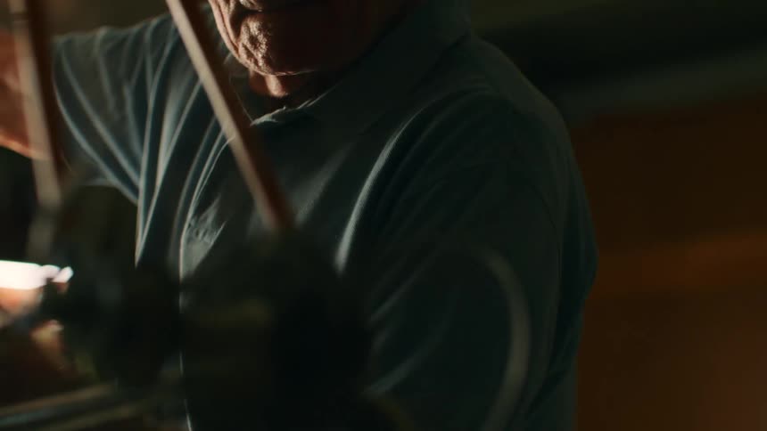 An elderly man with white hair wearing a blue polo shirt works on a bicycle in a dimly lit workshop