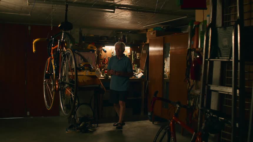 In a dimly lit garage an elderly man in a blue shirt is seen from behind working at a cluttered workbench before he turns around and approaches a bicycle hanging from the ceiling